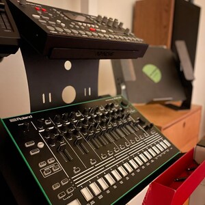 Op de afbeelding: Een zwarte en groene Roland TR-8S Rhythm Performer drummachine met een verscheidenheid aan knoppen, knoppen en pads. De drummachine staat op een witte standaard met een rode lade.