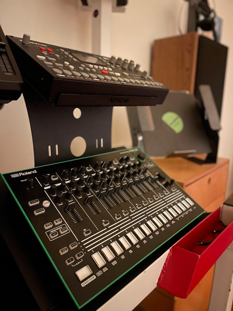 Op de afbeelding: Een zwarte en groene Roland TR-8S Rhythm Performer drummachine met een verscheidenheid aan knoppen, knoppen en pads. De drummachine staat op een witte standaard met een rode lade.
