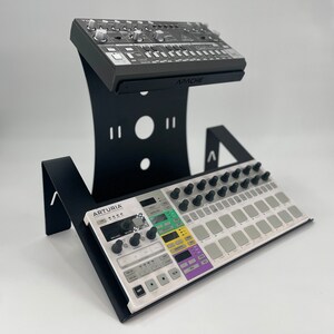 Op de afbeelding: Zwarte metalen standaard met twee niveaus voor het monteren van muziekapparatuur. Een zwarte en grijze synthesizer met het woord "APACHE" erop is gemonteerd op het bovenste niveau. Een witte en grijze synthesizer met het woord "ARTURIA" erop is gemonteerd op het onderste niveau.