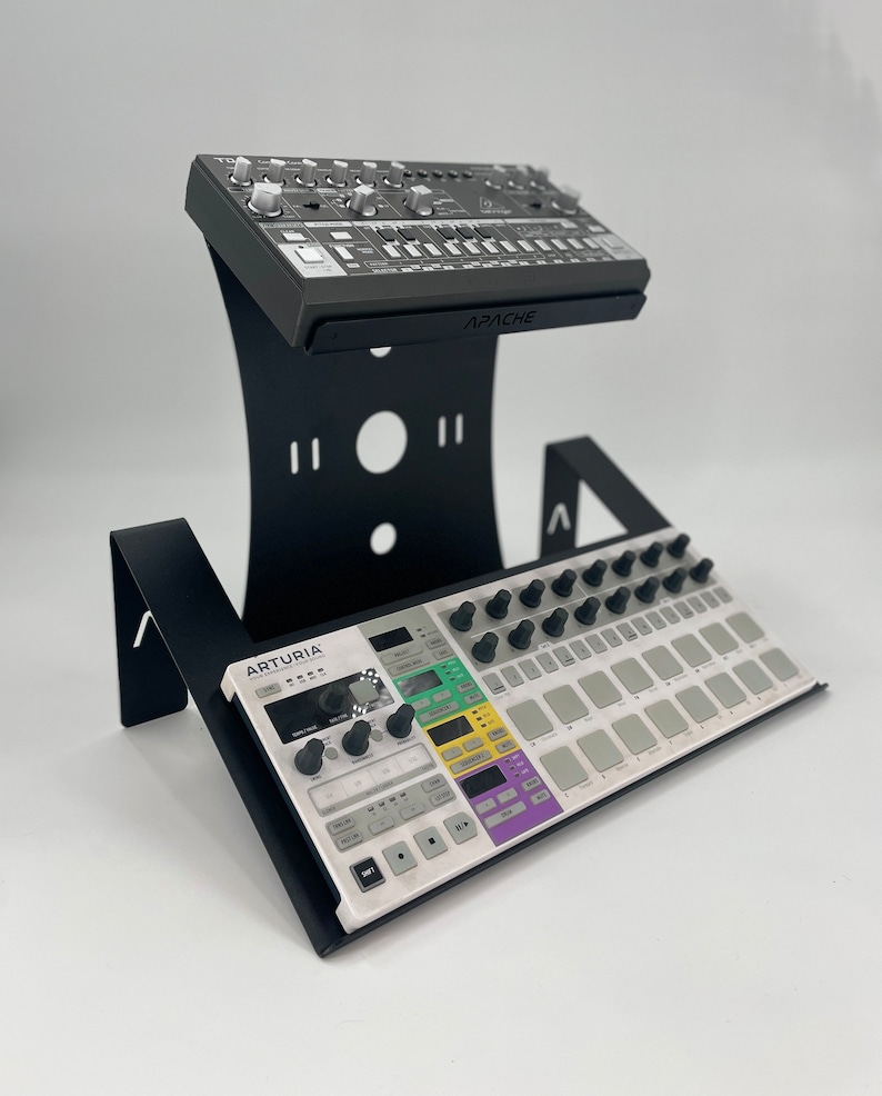 Op de afbeelding: Zwarte metalen standaard met twee niveaus voor het monteren van muziekapparatuur. Een zwarte en grijze synthesizer met het woord "APACHE" erop is gemonteerd op het bovenste niveau. Een witte en grijze synthesizer met het woord "ARTURIA" erop is gemonteerd op het onderste niveau.