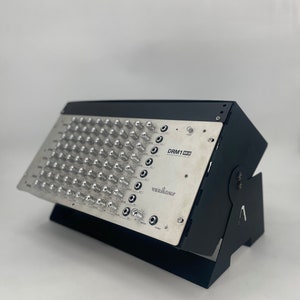 Peut inclure: Module de synthétiseur Verbos Electronics DRM1 MKII argenté et noir. Le module comporte de nombreux boutons et prises d'entrée, avec le texte "DRM1 MKII" et "Verbos Electronics" imprimé sur la face avant. Il est monté sur un support noir.