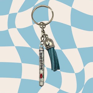 Puede incluir: Un llavero plateado con un pequeño tubo de caramelos Mentos y una borla turquesa. El tubo transparente de Mentos muestra los caramelos y tiene el logotipo de Mentos. El fondo es un patrón de cuadros azules y blancos.