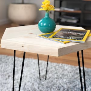 May include: A hexagonal wooden table with black hairpin legs. The table has a light wood finish and is topped with two magazines and a teal blue vase with yellow flowers.