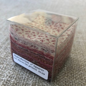 Puede incluir: Una caja de plástico transparente llena de 120 cuadrados de tela de 5 cm. Los cuadrados son de varios tonos de rojo y blanco con un patrón de anclas. La caja tiene una etiqueta que dice "Reproduction 2" Squares 120 Pieces (30 prints) $5.99".