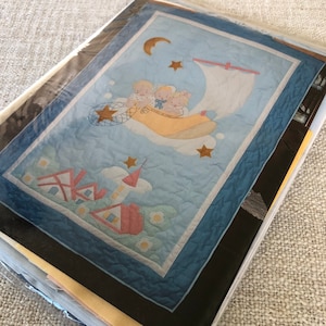 Puede incluir: Un edredón para bebé con un diseño caprichoso. Tres figuras querubinas están en un barco amarillo con forma de zapato navegando por un cielo azul con una luna creciente y estrellas. El edredón tiene un fondo azul claro con un borde y un diseño de casa y nube.