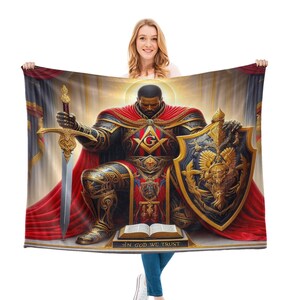Puede incluir: Una manta de forro polar roja, dorada y azul con una representaci&oacute;n de un hombre negro con armadura sosteniendo una espada y un escudo. El escudo tiene un s&iacute;mbolo mas&oacute;nico y el texto "In God We Trust".