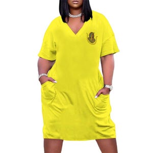 May include: A bright yellow V-neck dress with short sleeves and two front pockets. A small, circular emblem is on the left chest. The dress is a solid color and appears to be made of a soft fabric.