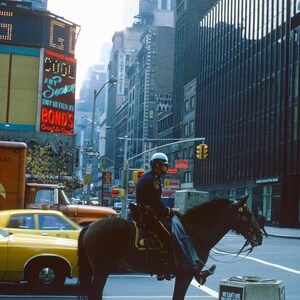 May include: A police officer on horseback rides through a busy city street in New York City. The horse is brown and the officer is wearing a blue uniform. There are tall buildings in the background and a yellow taxi cab in the foreground.