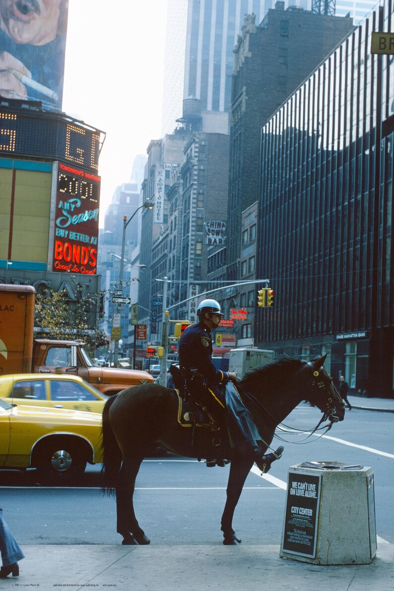 May include: A police officer on horseback rides through a busy city street. The horse is brown and black, and the officer is wearing a blue uniform and a helmet. The street is lined with buildings, and there are cars and pedestrians in the background. The officer is holding the reins in one hand and a whip in the other. There is a sign in the foreground that reads "We can't love alone. City Center."