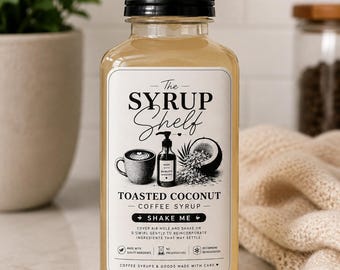 Könnte beinhalten: Eine durchsichtige Kunststoffflasche mit The Syrup Shelf Kaffeesirup mit geröstetem Kokosnussgeschmack und schwarzem Schraubverschluss. Das weiße Etikett zeigt schwarzen Text, eine Kaffeetasse und eine halbe Kokosnuss mit Raspeln.