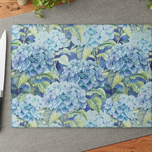 May include: Rectangular cutting board featuring a watercolor floral design of blue hydrangeas and green leaves on a dark blue background. A stainless steel knife is visible on the left.
