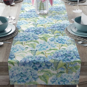 May include: A blue and green floral table runner with a watercolor design of hydrangeas. The runner is on a wooden table with place settings.