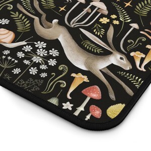 May include: A black mousepad featuring a whimsical illustration of a hare leaping through a forest scene. The design includes mushrooms, flowers, and foliage in shades of brown, red, yellow, and green. The mousepad has a black border.