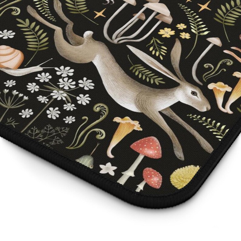May include: A black mousepad featuring a whimsical illustration of a hare leaping through a forest scene. The design includes mushrooms, flowers, and foliage in shades of brown, red, yellow, and green. The mousepad has a black border.