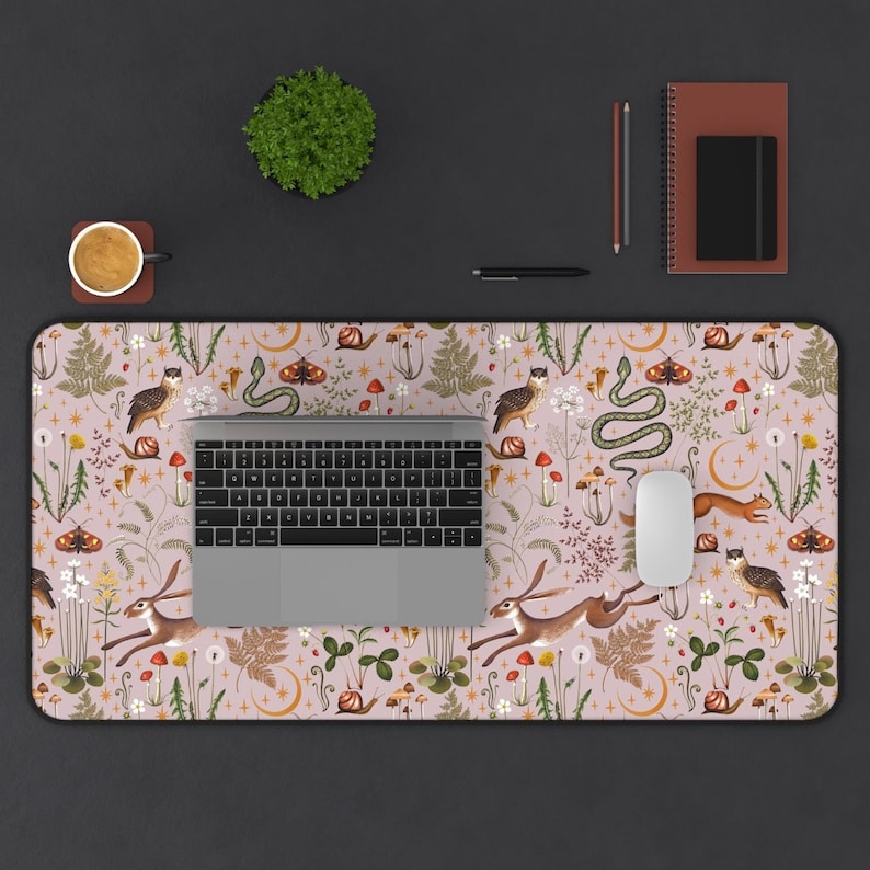 May include: A pink and white mouse pad with a whimsical woodland design featuring owls, butterflies, snakes, and hares. The design is surrounded by a border of green leaves and flowers. A laptop computer is sitting on the mouse pad.