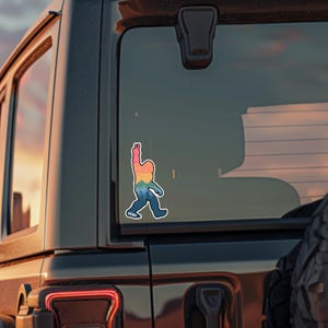 May include: A colorful sticker of a Bigfoot figure with an outstretched arm, featuring a rainbow gradient design. The sticker is affixed to a vehicle's rear window. The word "PEACE" is at the bottom of the sticker.