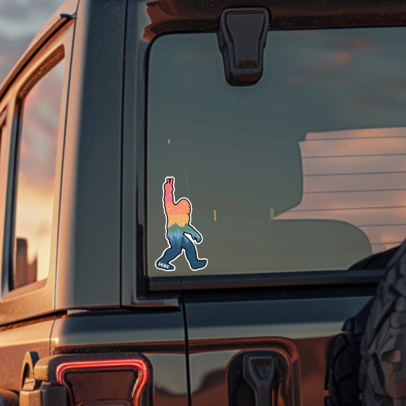 May include: A colorful sticker of a Bigfoot figure with an outstretched arm, featuring a rainbow gradient design. The sticker is affixed to a vehicle's rear window. The word "PEACE" is at the bottom of the sticker.