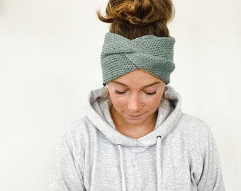 Op de afbeelding: Een saliegroene gebreide hoofdband met een gedraaid ontwerp. De hoofdband is gemaakt van een textuurrijk materiaal en wordt over het voorhoofd gedragen. De persoon op de foto draagt een grijze hoodie.