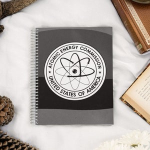 May include: A gray spiral notebook with a black and white logo of the Atomic Energy Commission. The logo features an atom with the text "Atomic Energy Commission United States of America".