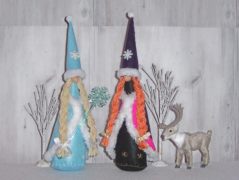 May include: Two felt Christmas ornaments, one in blue with a white snowflake and the other in purple with a white snowflake, stand in front of a white wooden background. A small felt reindeer stands in front of the ornaments.