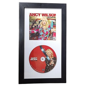 May include: A black framed photo with a white mount. The photo contains a CD and a CD cover. The CD cover features a woman with blonde hair and a red, white and black patterned shirt. The CD cover has the text "Nancy Wilson You and Me". The CD features a red background with the text "Nancy Wilson You and Me".