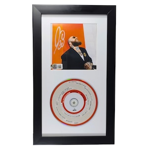 Pode incluir: Obra de arte emoldurada com uma fotografia de um homem de fato preto com uma assinatura e um CD. O CD tem círculos concêntricos vermelhos e brancos com títulos de músicas. A moldura é preta.