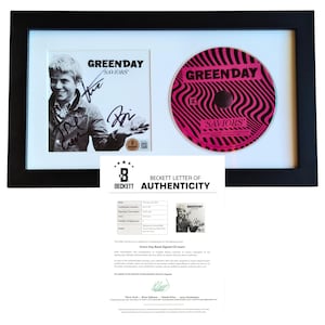 Pode incluir: Encarte e CD "Saviors" do Green Day emoldurados. O CD tem um design em espiral rosa e preto. O encarte apresenta uma foto em preto e branco de um membro da banda e autógrafos. Inclui uma carta de autenticidade Beckett.