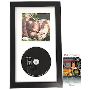 May include: A black framed photograph with a white mount and a CD. The photograph is of a man lying in grass. The CD is a music CD with the title "Love Goes" by Sam Smith. The photograph is signed by Sam Smith. The frame has a JSA authentication sticker.