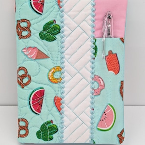 May include: A quilted fabric notebook cover in light mint green featuring a pattern of pretzels, cacti, watermelon slices, and ice cream. It includes a white diagonal quilted center panel, light blue trim, and a pink pocket holding a clear pen.