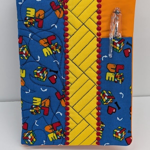 May include: A fabric notebook cover featuring a blue background with a repeating pattern of Rubik's cubes, red hearts, and the word LOVE. It includes a central yellow panel with diagonal stitching, red decorative trim, and an orange side pocket with a pen.