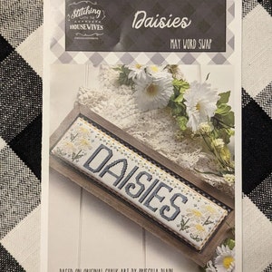 May include: A white and black word art design with the word "Daisies" in a black font on a white background. The design is framed in a wooden frame with a black and white checkered border. The design is based on original chalk art by Priscilla Blain.