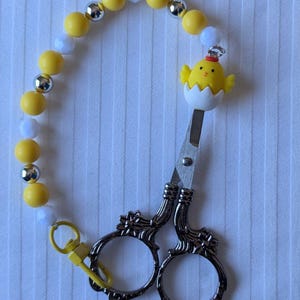 May include: Decorative embroidery scissors with a yellow beaded chain. The scissors have ornate handles and a charm of a yellow chick hatching from an egg. The chain includes yellow, white, and silver beads, and a yellow clasp.