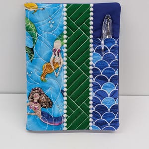 May include: A rectangular fabric notebook cover featuring a blue panel with mermaid illustrations, a central green quilted panel with a geometric pattern, and a blue scale-patterned pocket containing a clear plastic pen, all accented with white trim.