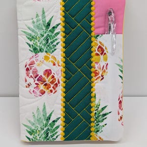 May include: A quilted fabric notebook cover featuring a white background with a colorful pineapple print. The cover includes a central dark green panel with a woven pattern, yellow pom-pom trim, and a solid pink fabric pocket holding a clear pen.