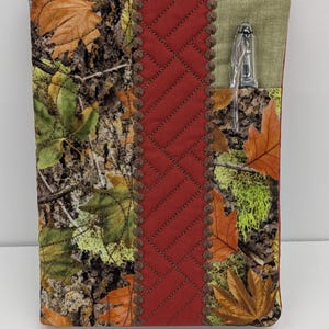 May include: A rectangular fabric book cover featuring a camouflage pattern of autumn leaves, tree bark, and moss. A central vertical band of solid dark red fabric with decorative stitching is flanked by a light green pocket holding a clear pen.