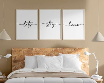 May include: Three framed prints with the text "let's stay home" in black cursive script on a white background. The prints are hanging on a beige wall above a white bed with a brown throw blanket.