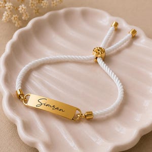 May include: A white cord bracelet featuring a gold-toned rectangular metal bar engraved with the name Simran in a black script font. The bracelet includes gold-toned metal end caps and a sliding adjustable gold-toned bead closure mechanism.