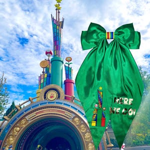 May include: A large, bright green satin bow with a rainbow stripe in the center. The bow has a gold graphic of a colorful tower and the text "HERE WE GO!" on one of the tails. The background features a colorful building.