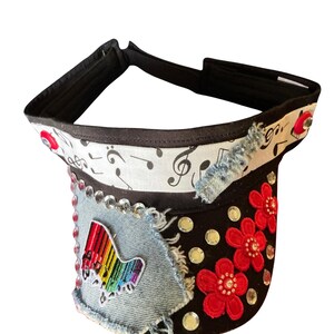 Può includere: Una visiera nera con una fascia bianca decorata da note musicali nere, una toppa in denim azzurro sfrangiato con un piano arcobaleno, tre applicazioni floreali rosse ricamate e strass trasparenti sparsi sulla parte anteriore.