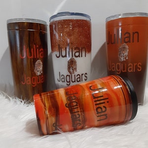 May include: Four insulated tumblers with lids. The tumblers feature the name "Julian" and the word "Jaguars" in red, along with a jaguar logo. The tumblers have various designs, including wood grain, glitter, and an orange and brown swirl.