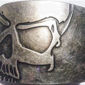 May include: A silver metal belt buckle with a skull design. The skull is detailed with a prominent eye socket and teeth.