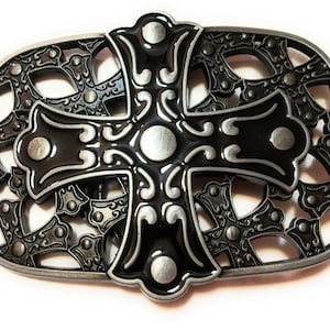 Puede incluir: Una hebilla de cinturón plateada con un diseño de cruz negra. La cruz está rodeada por un borde decorativo de cruces más pequeñas.