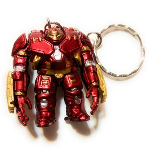 May include: A red and gold metal keychain featuring a Hulkbuster Iron Man figure. The figure is standing with its arms outstretched and has a detailed design.