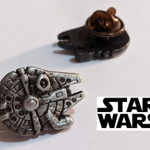 Könnte beinhalten: Zwei silberne Star Wars-Anstecknadeln aus Metall. Eine Nadel zeigt eine detaillierte Darstellung des Raumschiffs Millennium Falcon. Die andere Nadel ist eine Silhouette eines TIE-Jägers.