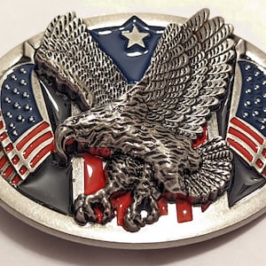 Pode incluir: Uma fivela de cinto de prata com um design de águia careca. A águia está segurando duas bandeiras americanas em suas garras. As bandeiras são vermelhas, brancas e azuis.