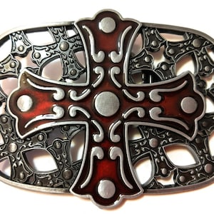 Puede incluir: Una hebilla de cinturón plateada con un diseño de cruz roja. La cruz está rodeada por un borde decorativo de cruces más pequeñas.