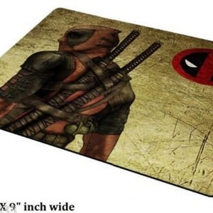 May include: A brown and yellow mousepad with a graphic of Deadpool, a superhero wearing a red and black suit, holding two swords. The background is a distressed, textured design. The mousepad also features a red and black Deadpool logo.