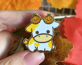 Puede incluir: Un pin de esmalte dorado con una vaca de dibujos animados blanca con manchas amarillas que lleva dos sombreros de calabaza naranja. El pin está en una mano que sostiene una hoja marrón y naranja.