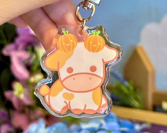 May include: A clear acrylic keychain featuring a cartoon cow with orange pumpkin accents on its head. The cow is white with orange spots and has a pink nose and hooves.