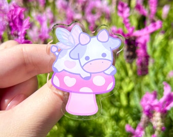 May include: A pink and white cow with fairy wings sits on a pink mushroom with white polka dots. The cow has a pink heart on its side and a pink nose.